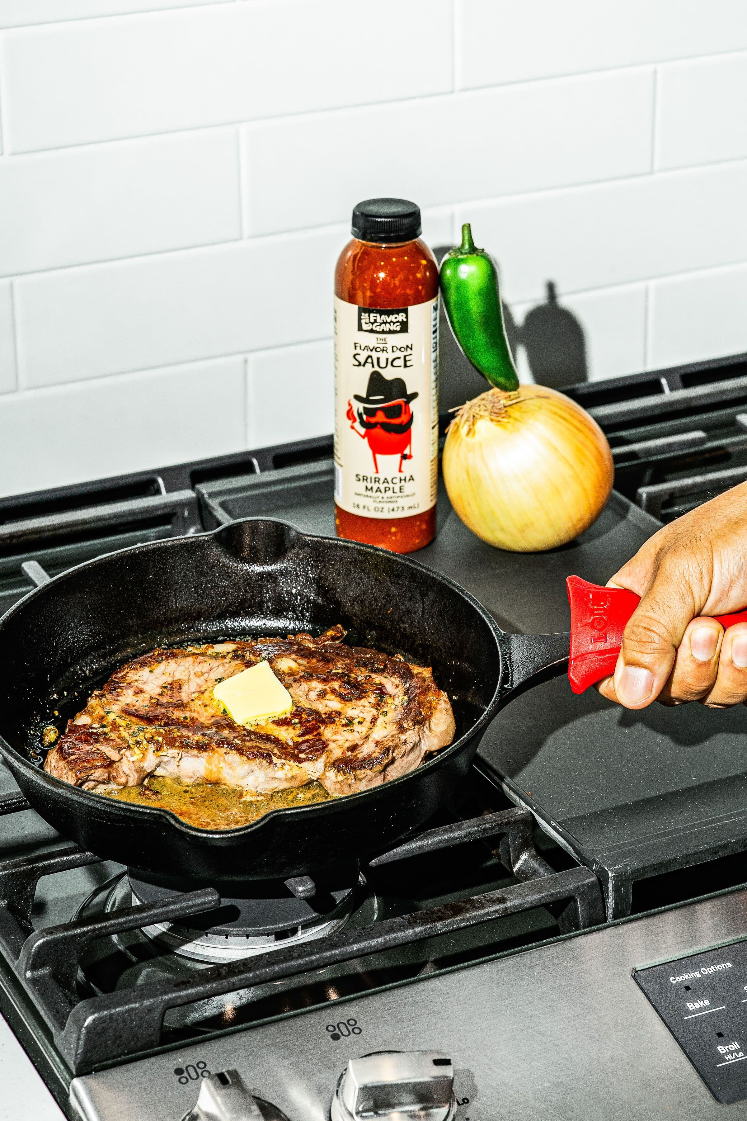 Cast iron steak cooking with butter on a stovetop beside The Flavor Gang Flavor Don Sriracha Maple sauce, onion, and jalapeño.